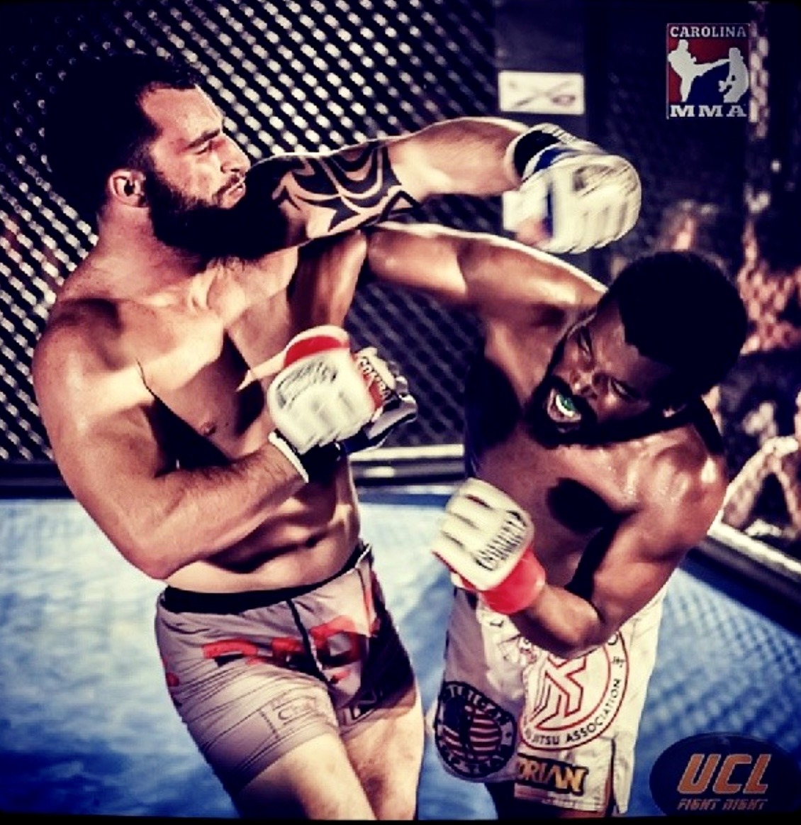 Two fighters exchanging strikes inside a cage during a mixed martial arts bout, the bearded fighter at left in grey and red fight shorts throwing a punch