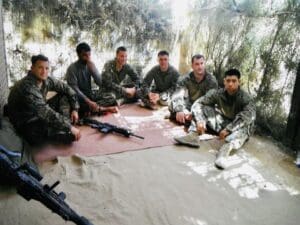 Six Marines in camouflage utilities sitting on the ground against a mud-brick wall with rifles laid beside them, eating a meal