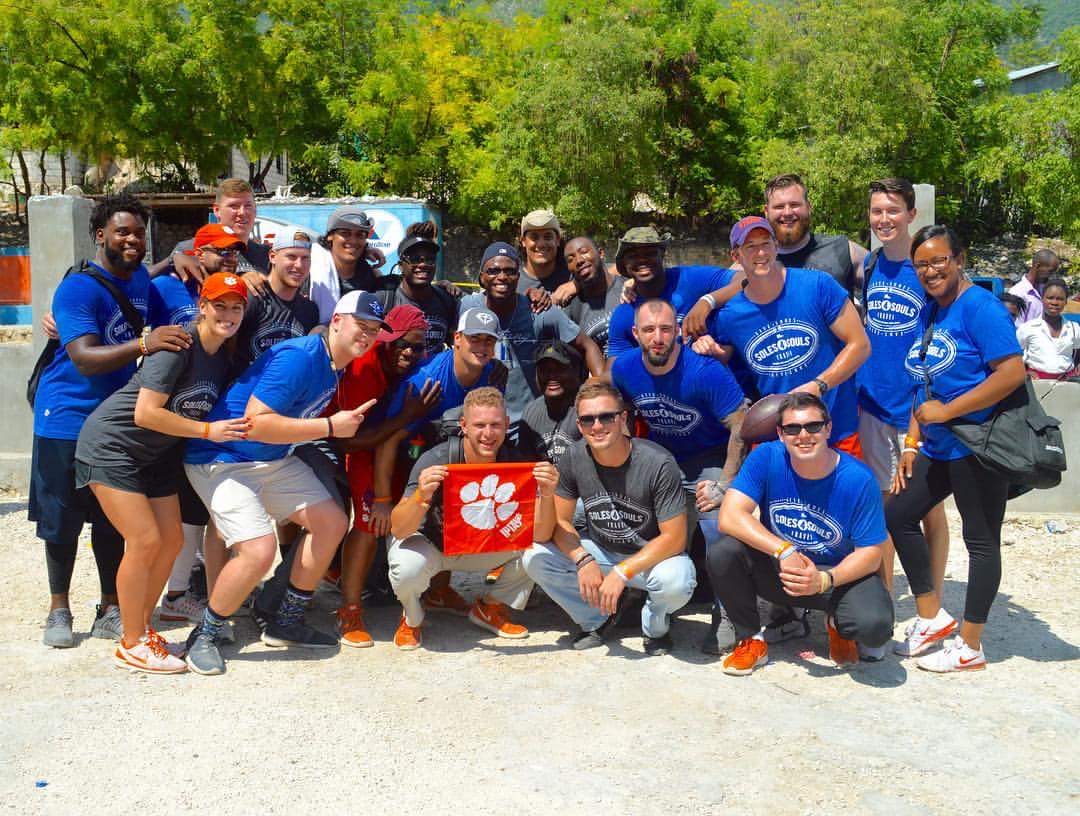 Large volunteer team in blue Soles4Souls shirts standing and kneeling together outdoors on a dirt road, several holding an orange banner