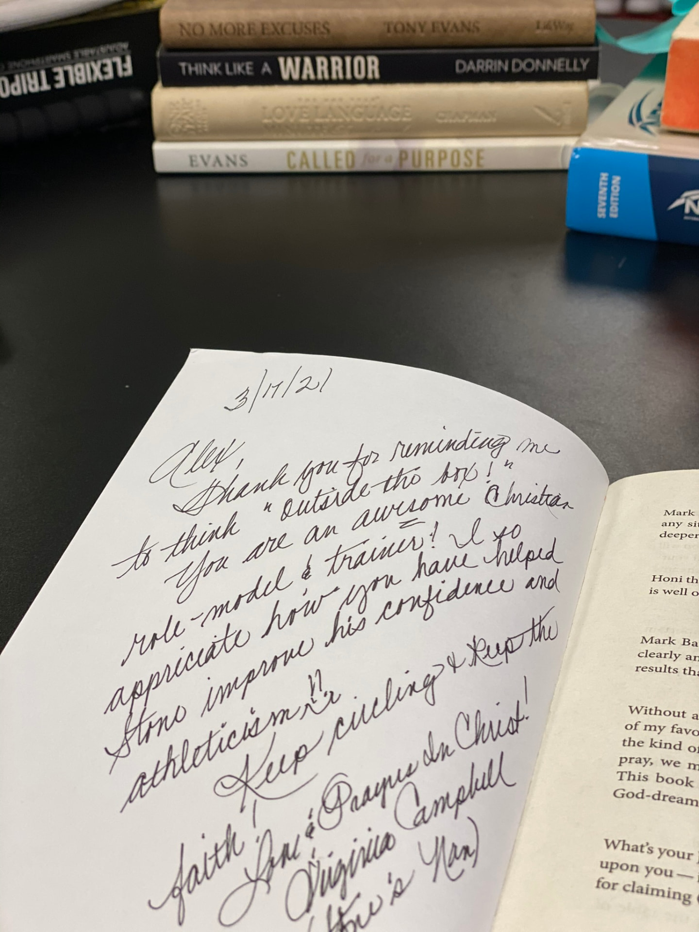 A handwritten inscription in cursive ink on the opening page of a book, dated 3/17/21 and addressed to Alex, thanking him as a Christian role model and trainer, signed Virginia Campbell, with a stack of books on the desk behind it