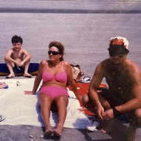 Faded beach snapshot of a woman in a pink swimsuit and sunglasses seated on a towel, a boy sitting on the sand behind her, and a man in a cap and sunglasses beside her