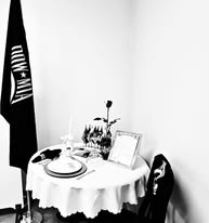 Black and white photograph of a small round table set for one in a corner of a room, with a single rose in a vase, an inverted glass, a candle, a framed photograph, an empty chair, and a black flag on a stand beside it