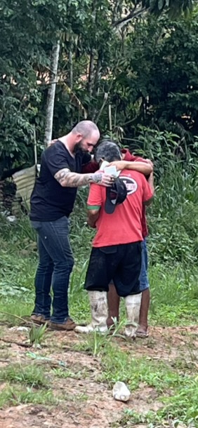 Two men praying with a third man in a red shirt, hands on his shoulders, standing in a grassy clearing at the treeline