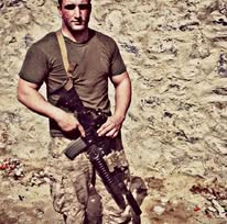 A Marine in a tan T-shirt and camouflage trousers standing beside a rough stone and mud wall, holding a rifle on a sling