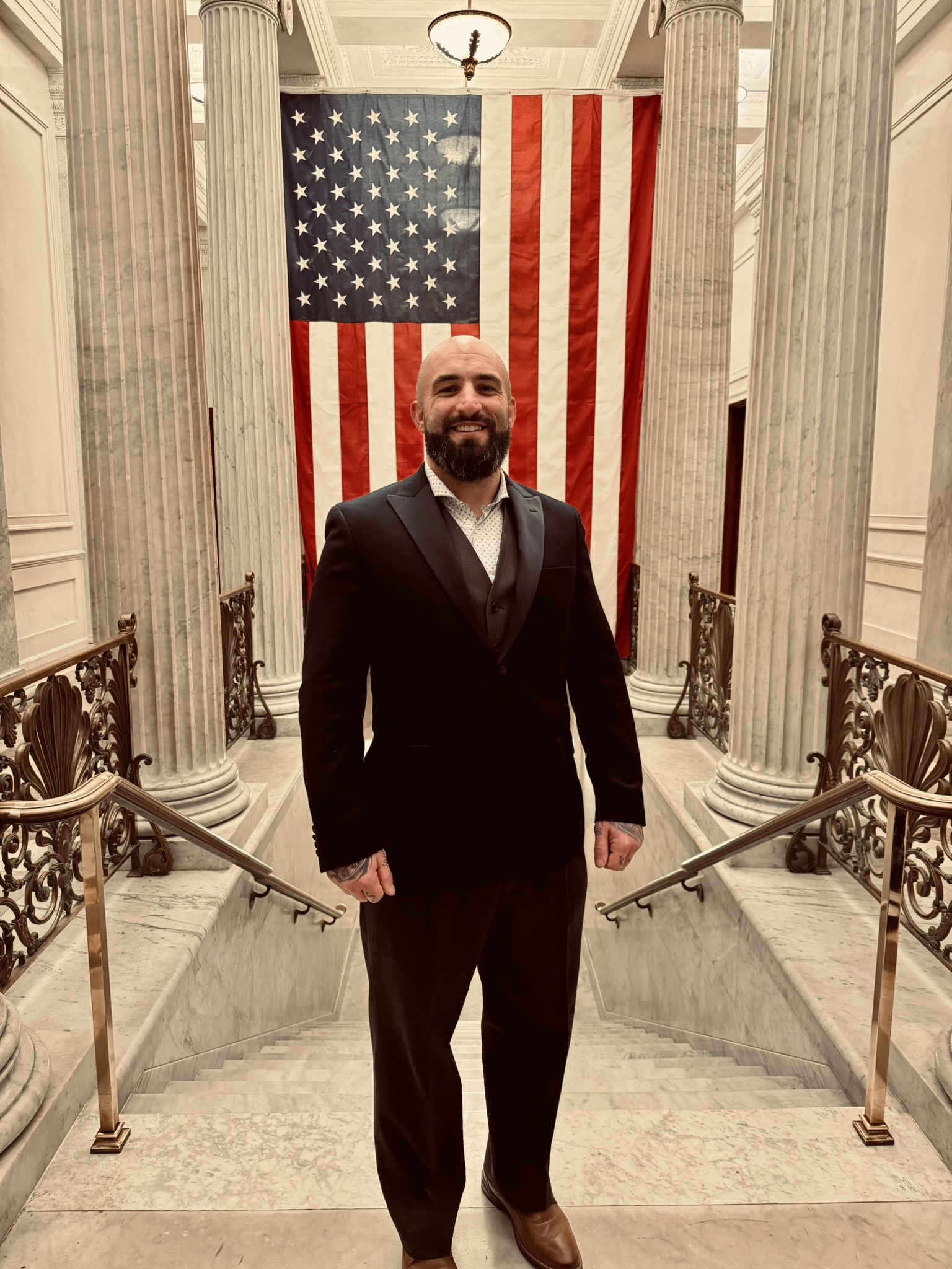 Johnathan Alexander Dalton in a dark three-piece suit, standing on a white marble staircase before a large hanging American flag