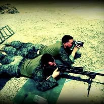 Two Marines lying prone in the dirt in camouflage utilities, one sighting through an optic and one behind a rifle on a bipod