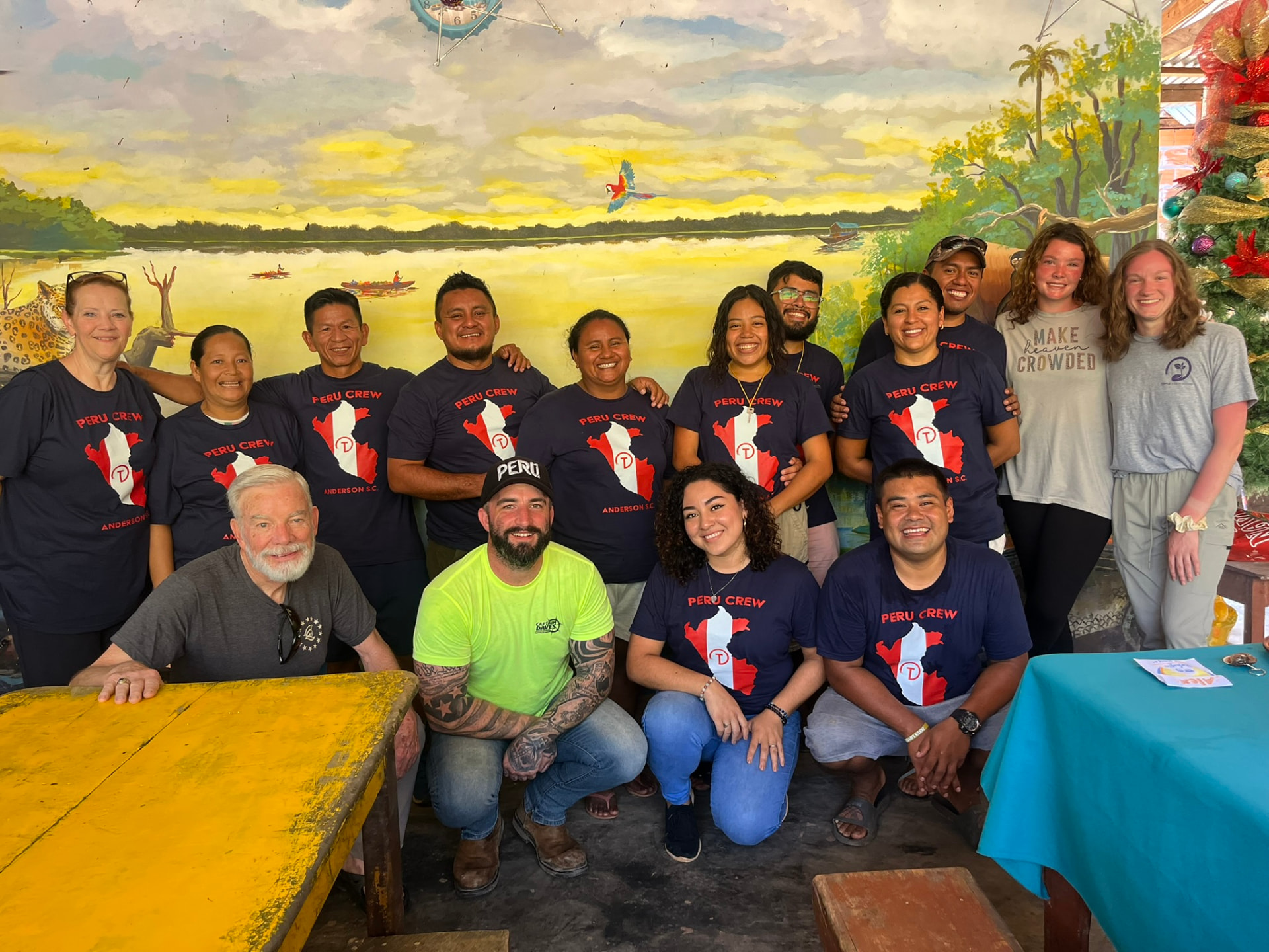 Mission team of about fourteen people in navy Peru Crew shirts, standing and kneeling together in front of a painted river mural