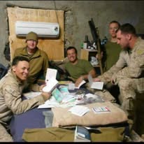 Five service members gathered around a table in a plywood-walled room, opening letters and packages, with a wall-mounted air conditioning unit behind them