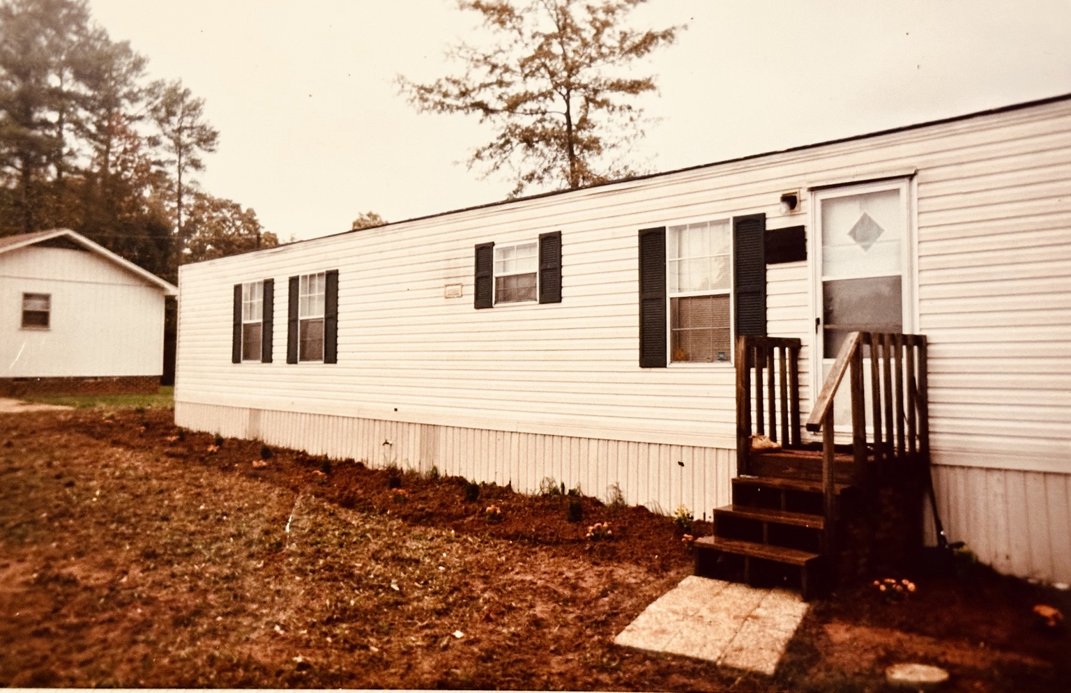 Faded snapshot of a white single-wide trailer home with dark shutters and wooden front steps, bare dirt yard and pine trees behind it