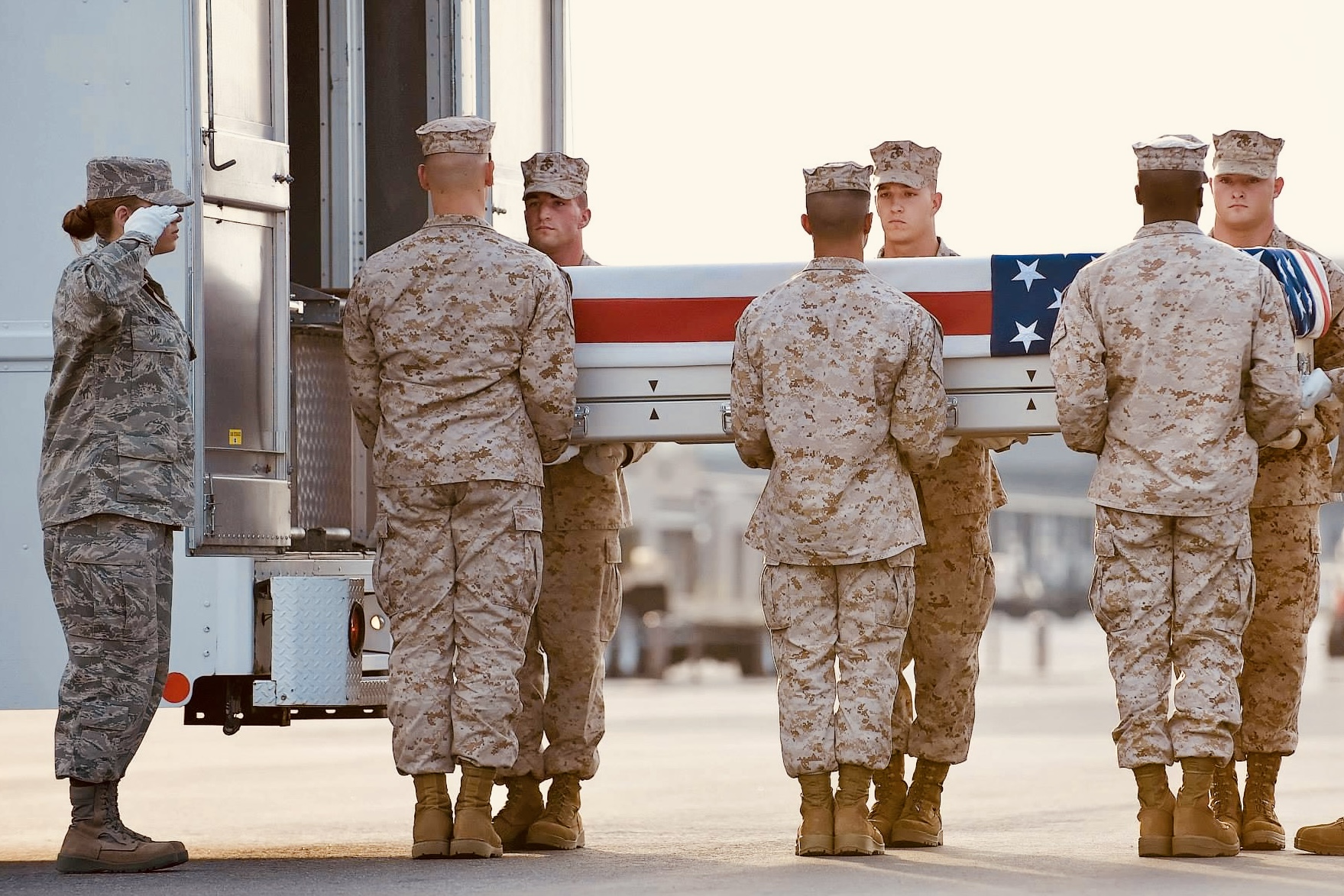 A carry team of service members in desert camouflage utilities carrying a flag-draped transfer case on a flight line while another service member renders a hand salute