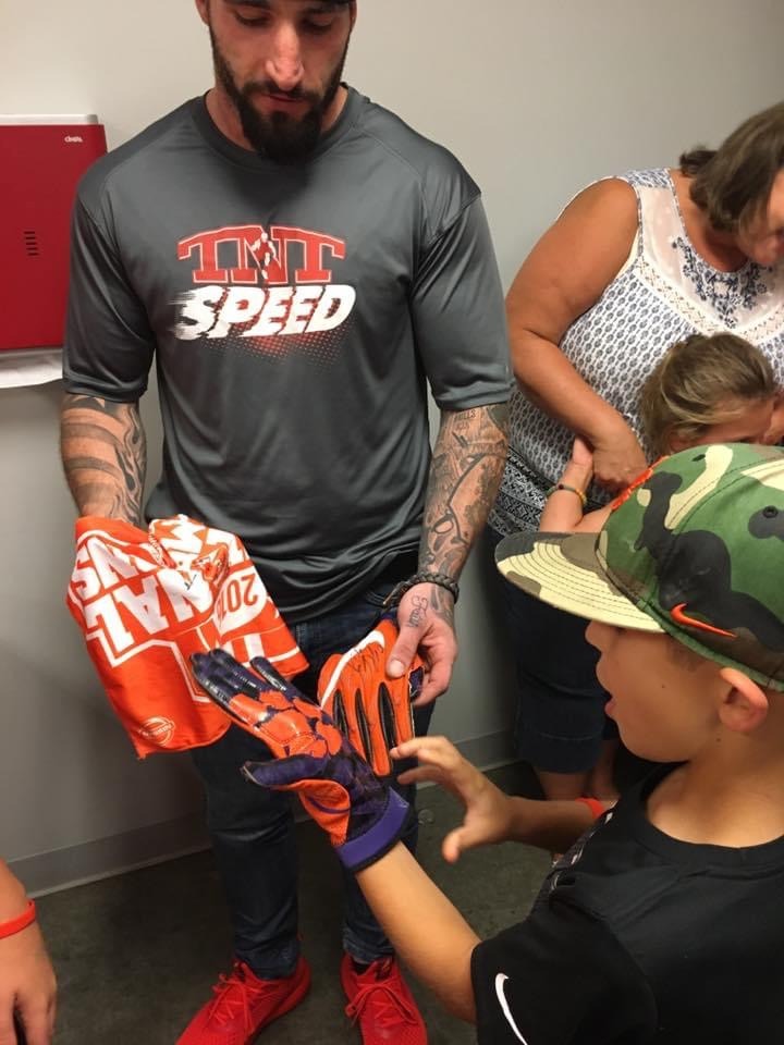 A man in a coaching shirt kneeling to fit a pair of gloves onto a young participant at a youth sports event, with other attendees nearby