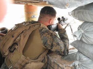 A Marine in a plate carrier and pack kneeling behind a rifle in a fighting position built from sandbags, sighting through a scope out of a firing port