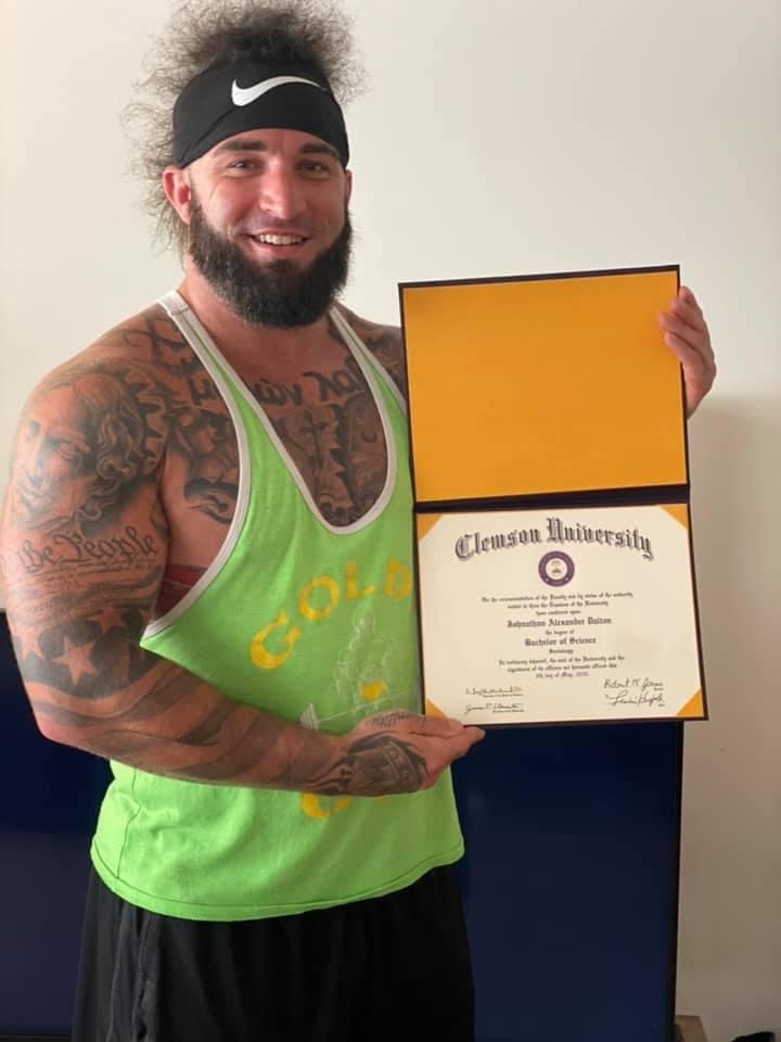 The author, bearded and in a green tank top, holding open a Clemson University diploma in its folder and smiling