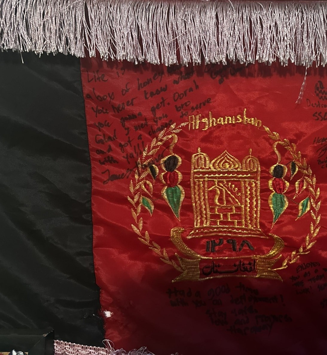 Detail of an Afghanistan flag with gold fringe hanging on a wall, centered on the embroidered gold national emblem on the red panel, with handwritten messages and signatures in black ink around it