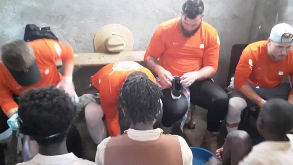 Volunteers in orange shirts seated inside a concrete-walled room, fitting new shoes onto children who are seated in front of them