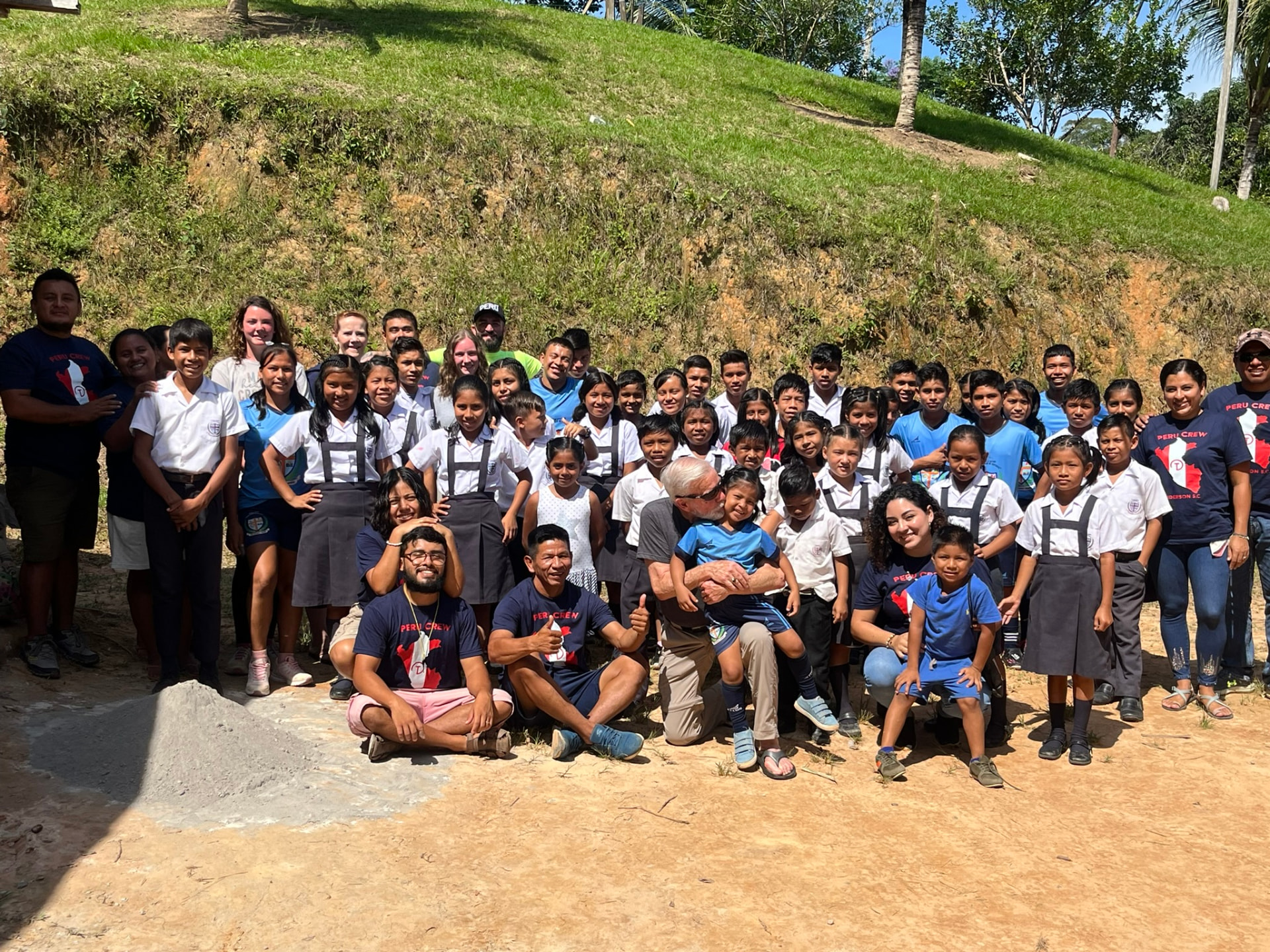 Large group of schoolchildren in white and grey uniforms gathered outdoors on a dirt slope with visiting mission team members