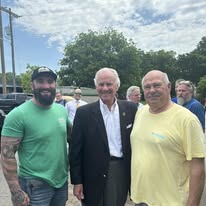 The author in a green shirt standing outdoors with Governor Henry McMaster in a dark suit and tie and Bill Biggs in a yellow shirt, with other people and trees behind them