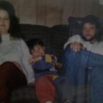 Faded indoor snapshot of a woman on a couch beside a small child in a red and blue outfit, with a second young person in a dark jacket and jeans sitting at the other end